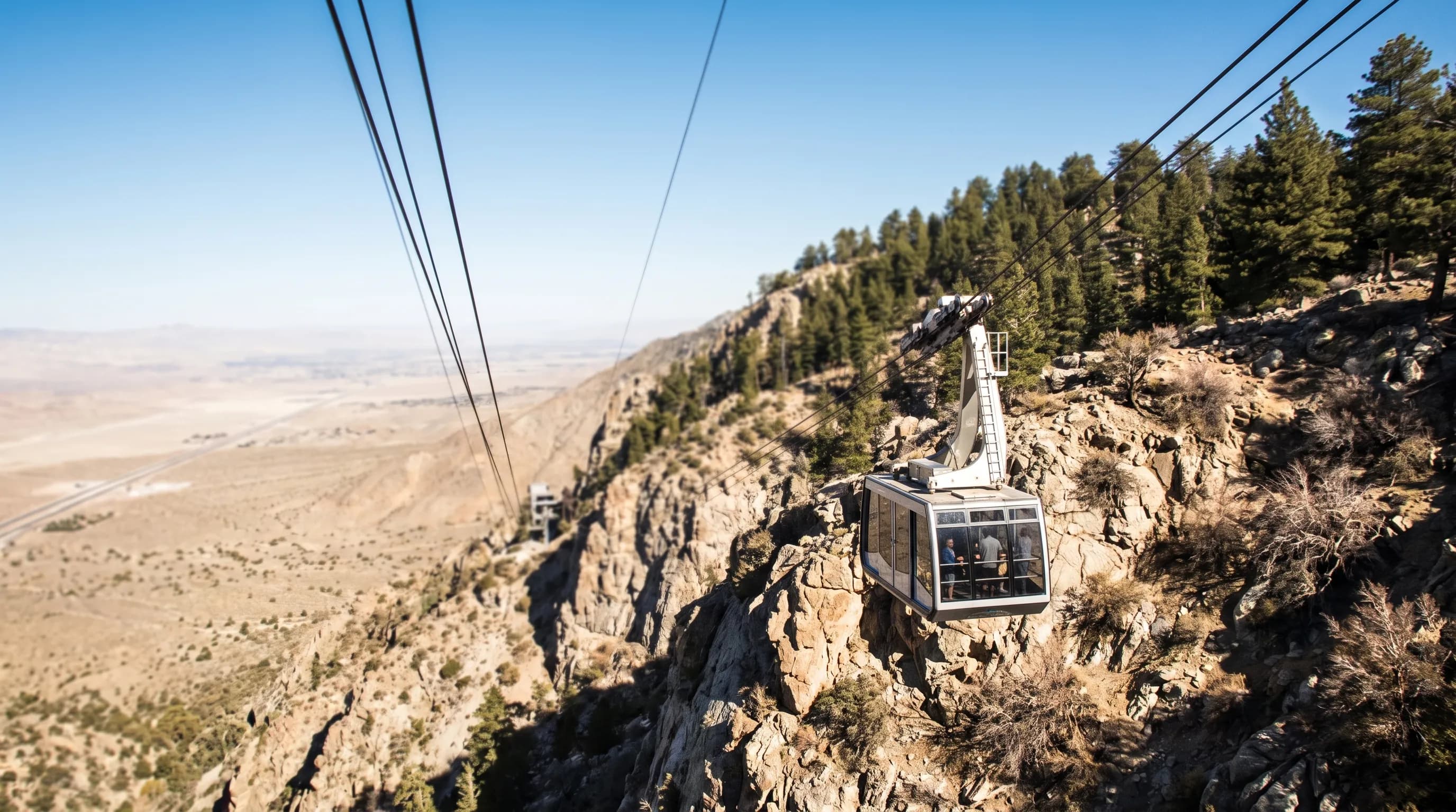 An aerial tram car climbing a steep, rocky mountainside from the desert floor toward pine-covered peaks under a clear sky